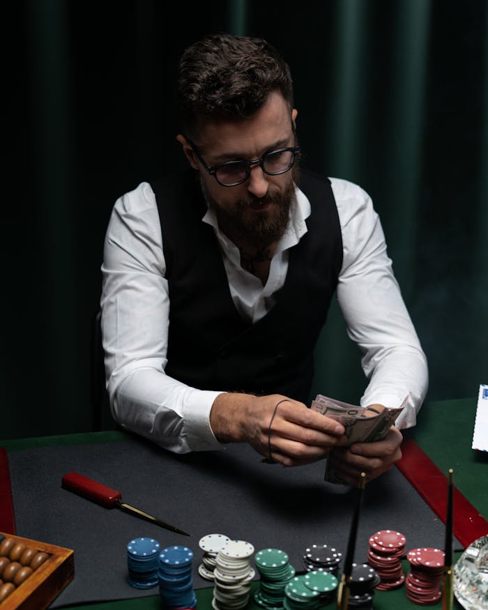 A focused bearded man counting money at a poker table with chips and cards.