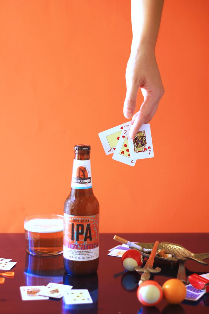 A vibrant still life featuring a bottle of IPA, playing cards, and billiard balls on an orange backdrop.